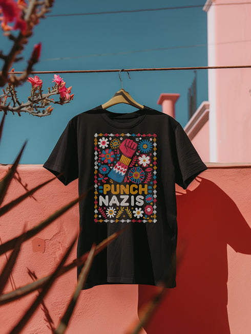 Black t-shirt featuring an embroidered-style design with the phrase "PUNCH NAZIS" in bold, colorful lettering. Above the text, a raised red fist is illustrated, symbolizing resistance, and is surrounded by a vibrant folk-art-inspired floral border with bright flowers, leaves, and geometric patterns in red, yellow, blue, and white.